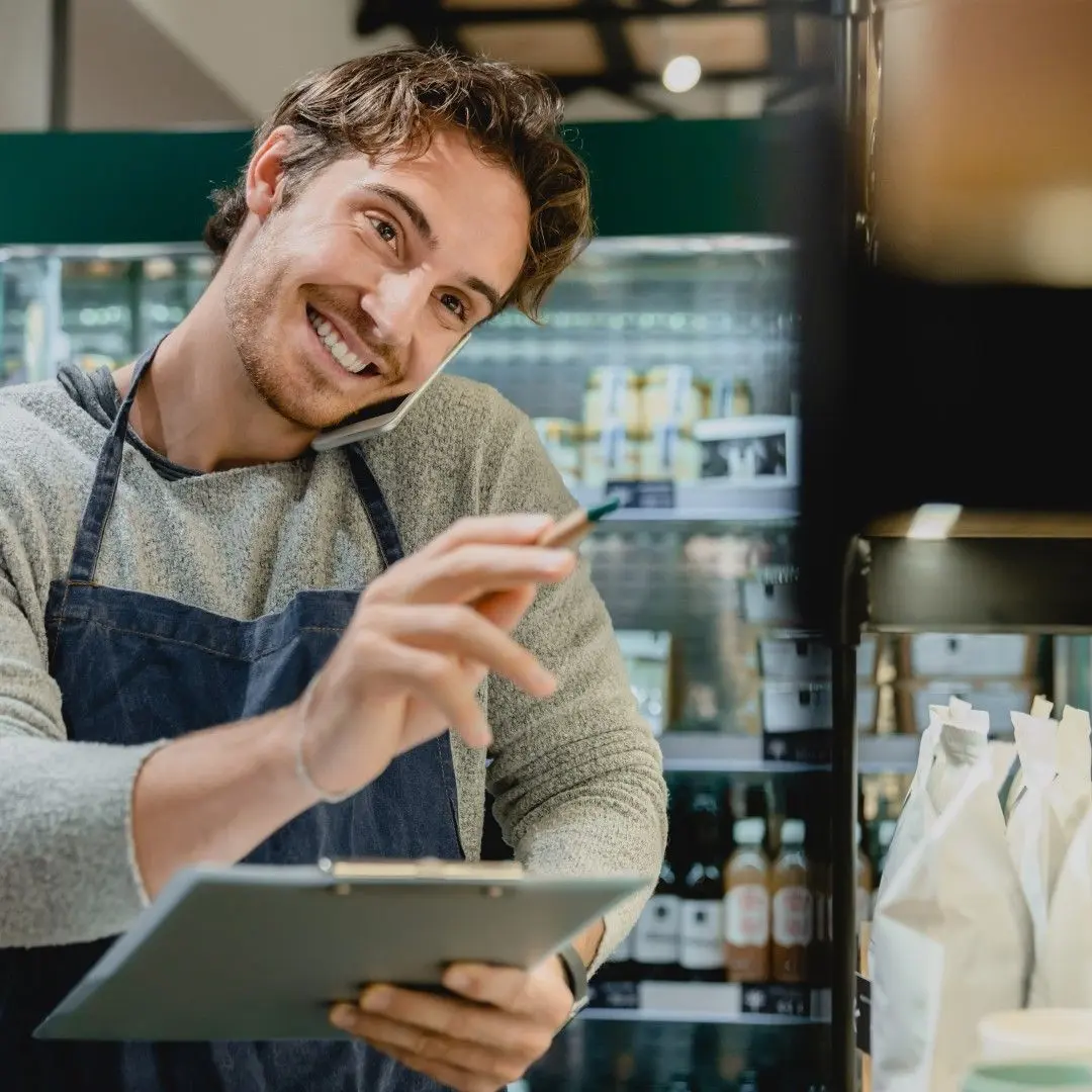 Un jeune vendeur souriant en tablier gère les stocks et téléphone dans un magasin.