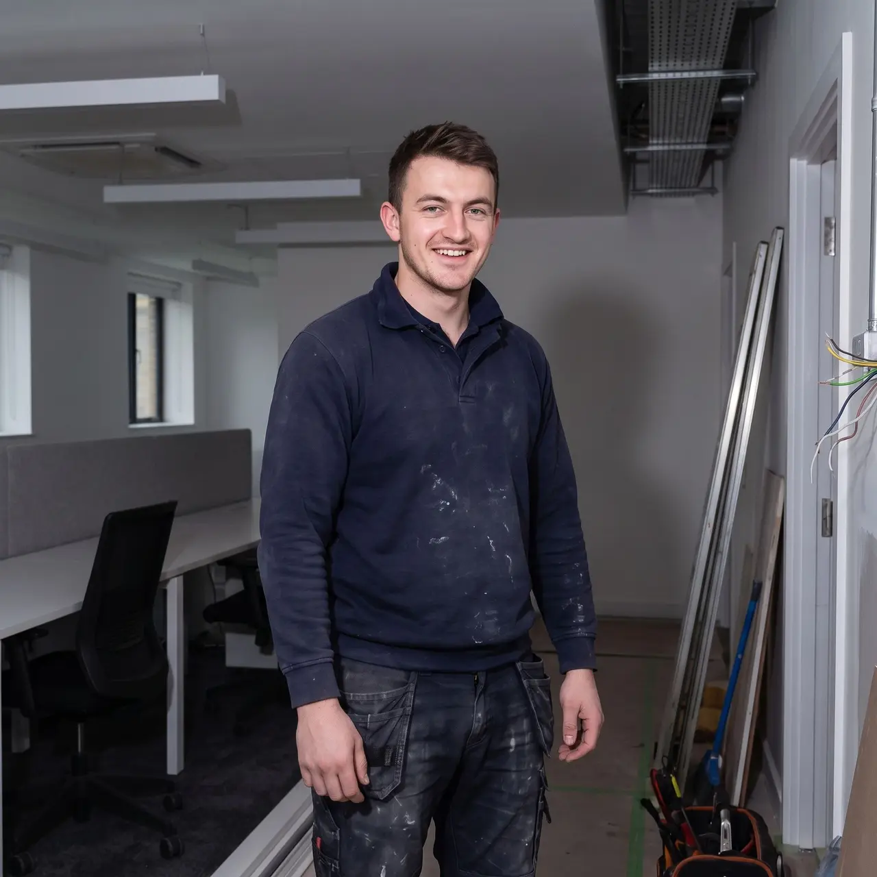 Un artisan en tenue de chantier sourit debout dans un bureau en rénovation.