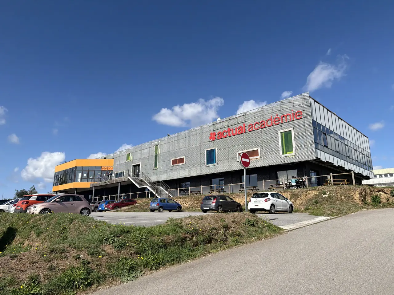 Façade du bâtiment de l'Actual Académie avec des voitures stationnées sur le parking devant.