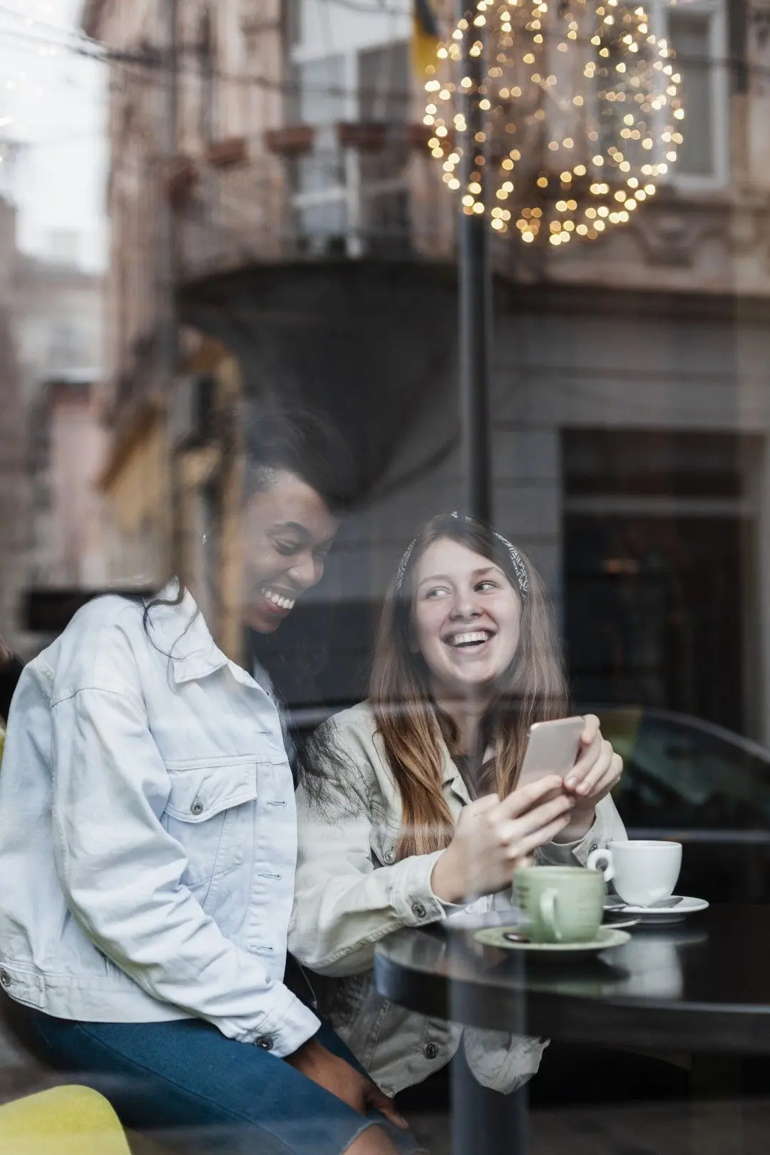 Deux femmes souriantes partagent un moment convivial dans un café en regardant un smartphone.