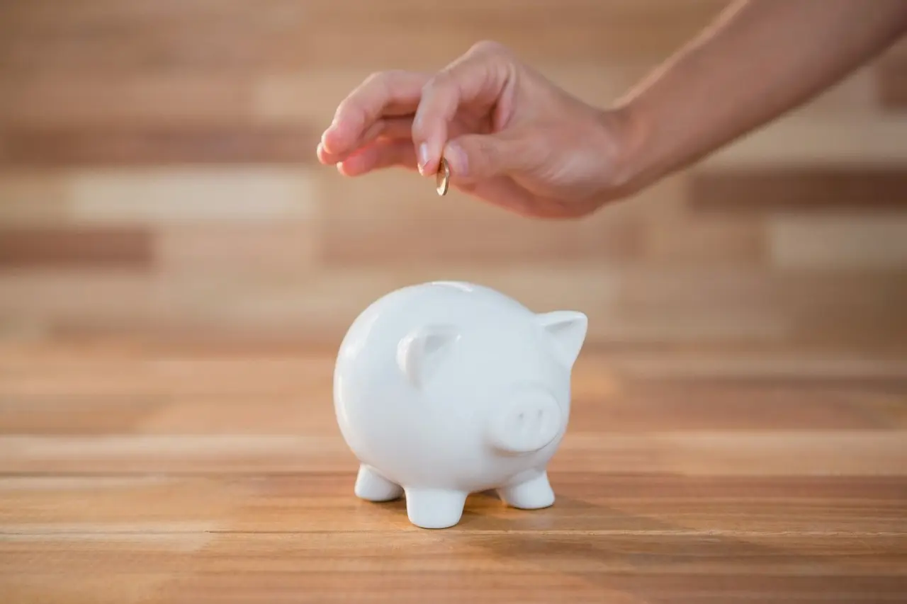 Une main glisse une pièce de monnaie dans une tirelire cochon blanche posée sur une table en bois.