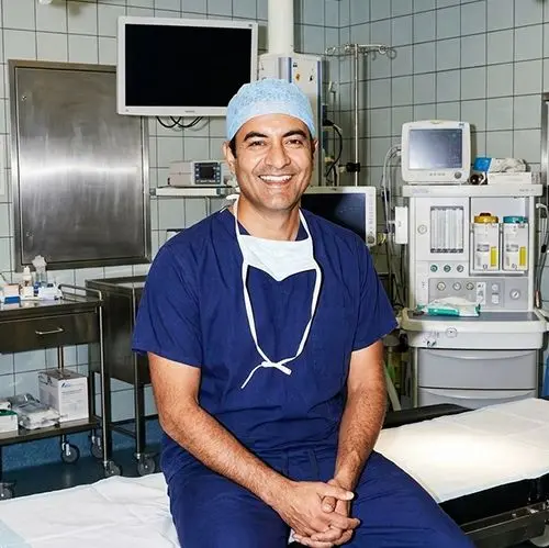 Un chirurgien souriant en tenue bleue assis sur une table d'opération dans un bloc chirurgical.
