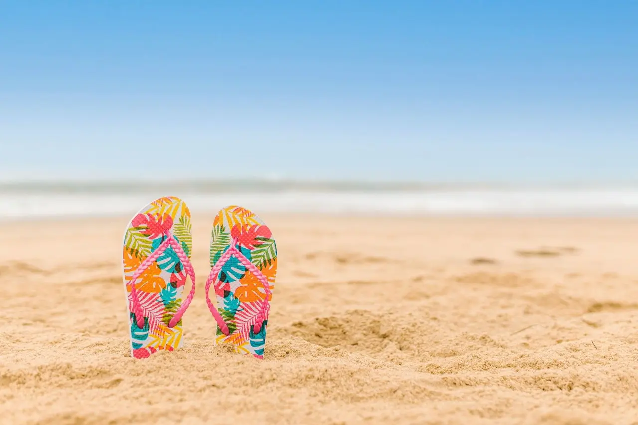 Paire de tongs colorées plantées dans le sable d’une plage face à la mer.