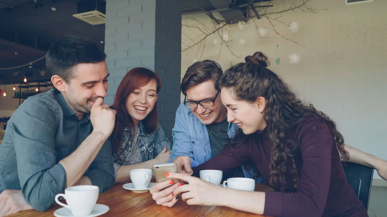 Quatre jeunes proches s’entraident et sourient autour d’un smartphone dans un café.