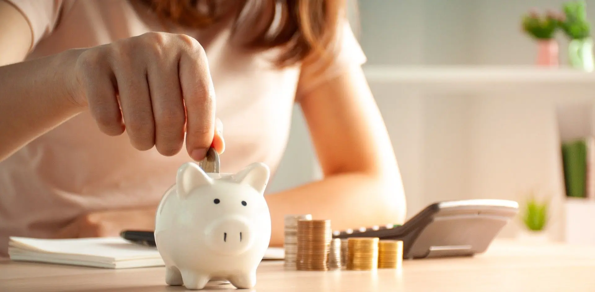 Une personne dépose une pièce de monnaie dans une tirelire cochon blanche à côté de piles de pièces.