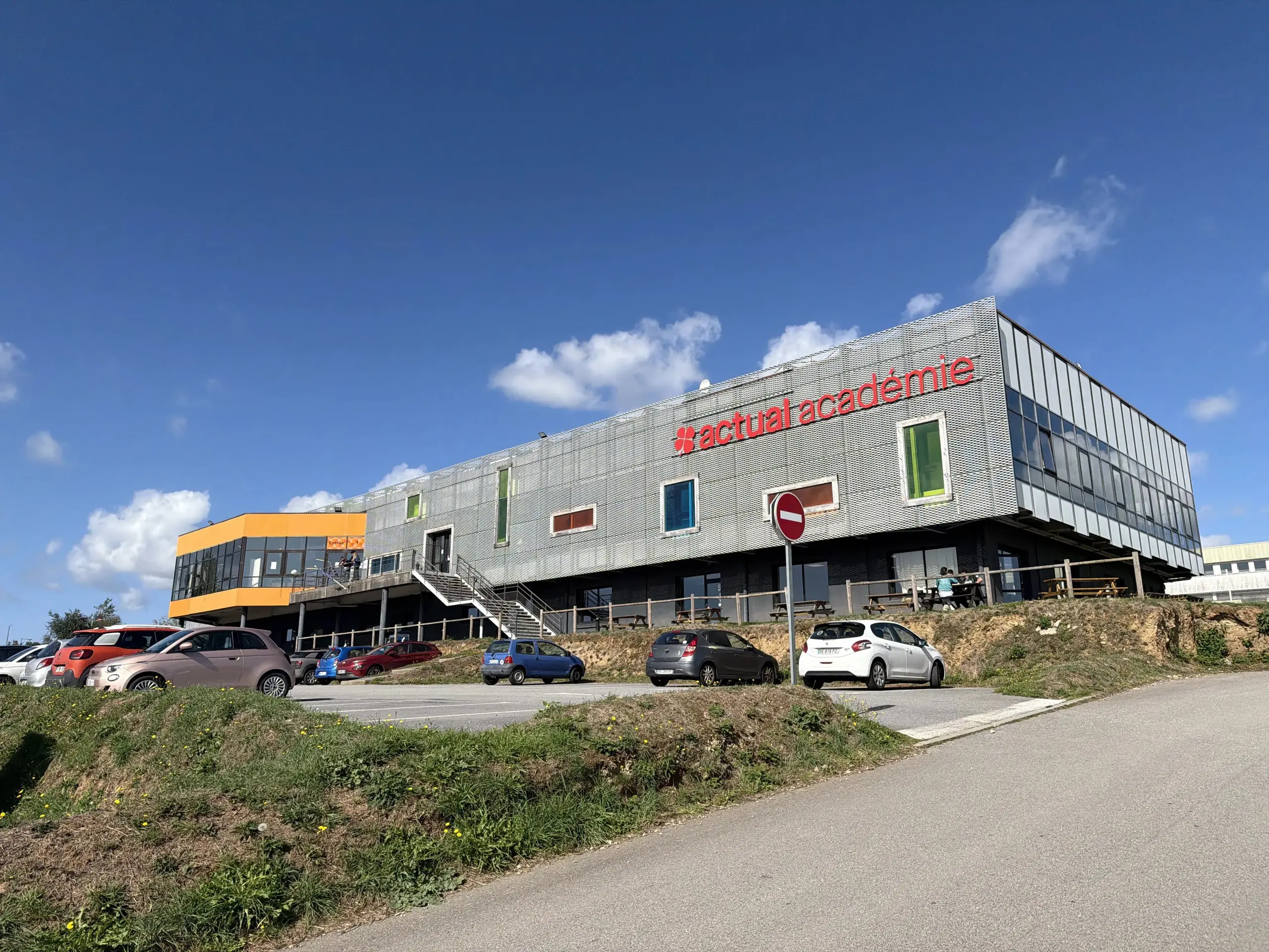 Façade du bâtiment de l'Actual Académie avec des voitures stationnées sur le parking devant.