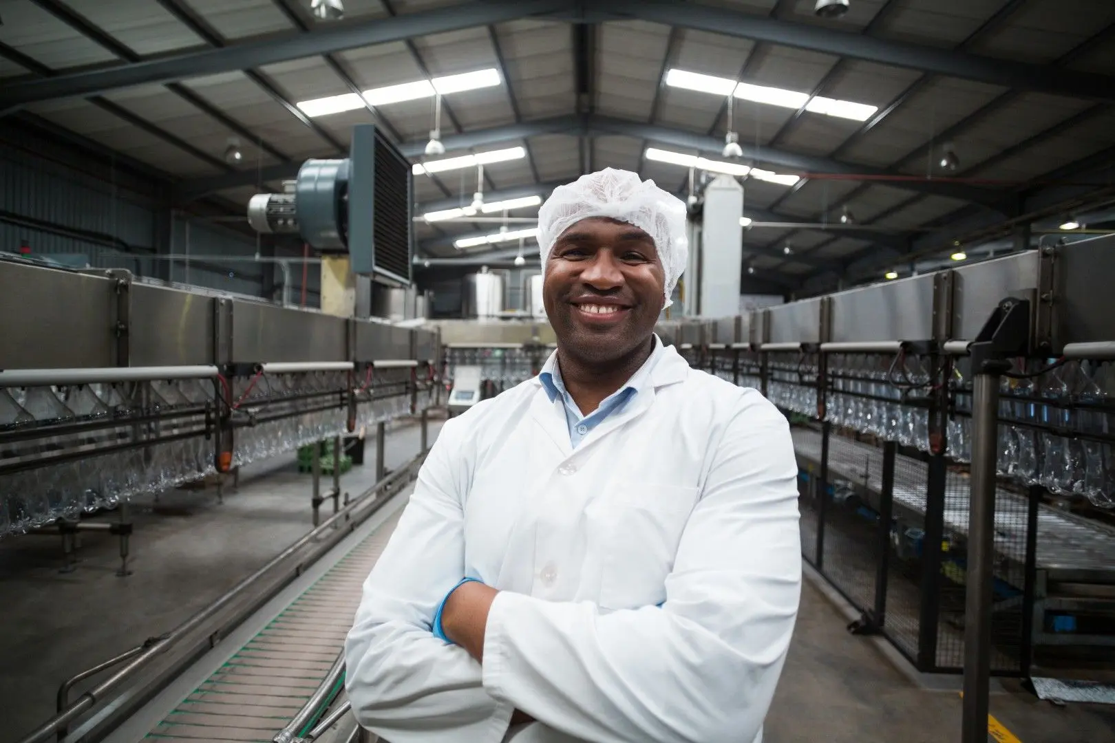 Un agent de conditionnement souriant, en blouse et charlotte, devant une ligne de production.