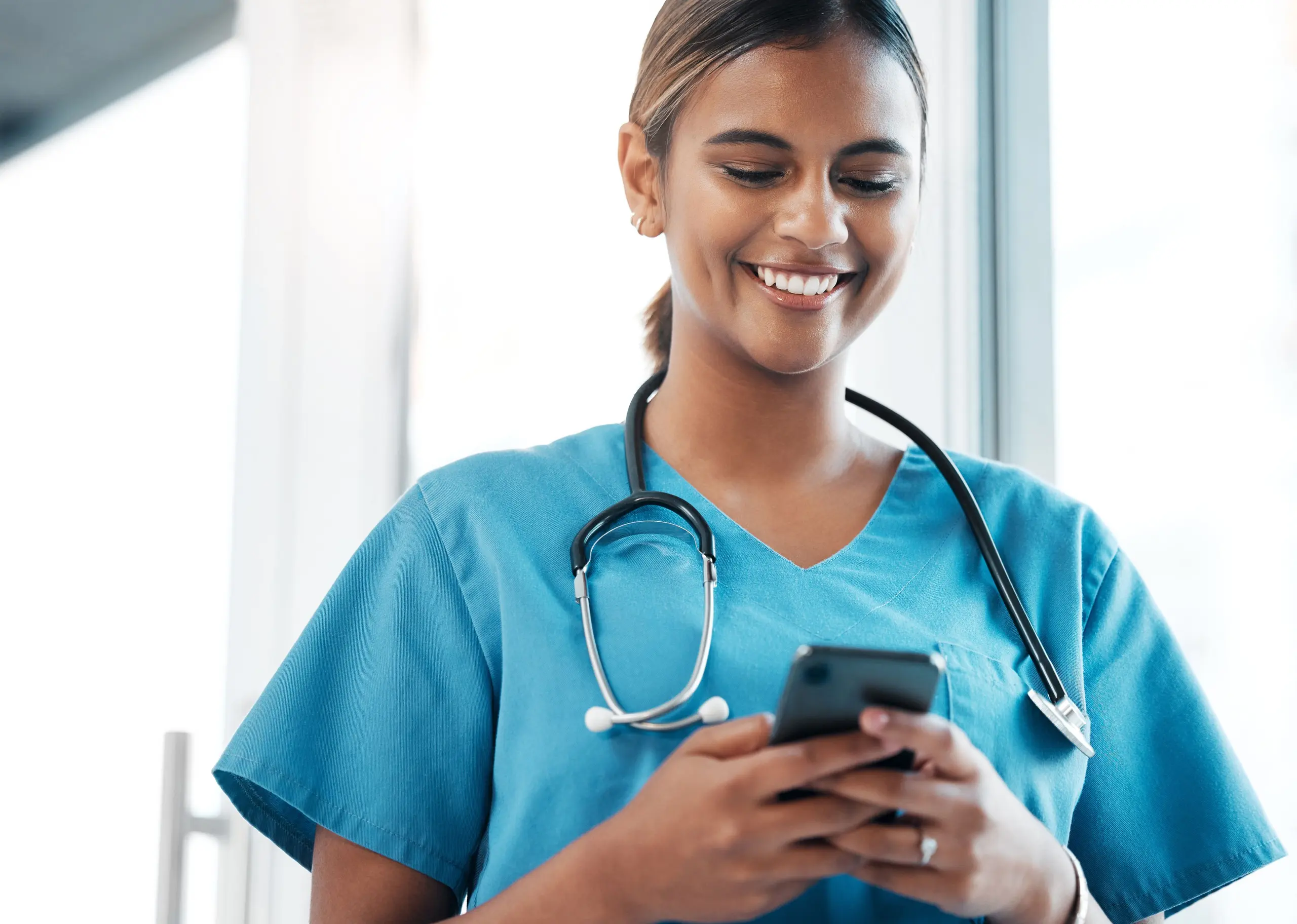 Une infirmière en blouse bleue avec un stéthoscope consulte son téléphone portable en souriant.