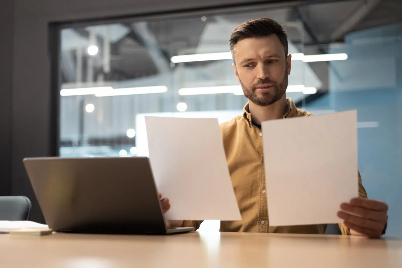 Un homme assis à son bureau compare deux documents papier devant son ordinateur portable.