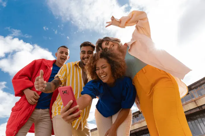 Quatre jeunes collaborateurs souriants prennent un selfie en extérieur sous un ciel bleu.