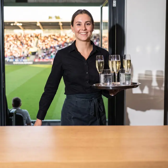 Une serveuse souriante tient un plateau de boissons lors d'un événement dans un stade de football.