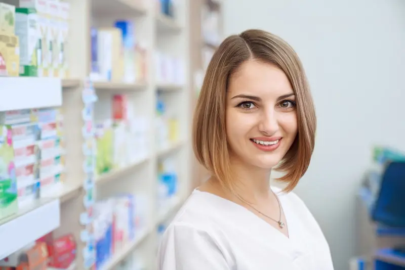 Une préparatrice en pharmacie souriante en blouse blanche devant des étagères de médicaments.